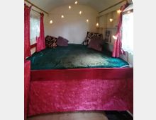 Innenraum eines kleinen Wohnwagens mit einer gemütlichen Schlafnische, die ein großes Bett mit dunkelgrüner Decke und mehreren Kissen umfasst. Der Raum ist dekoriert mit roten Vorhängen, Spitzenvorhängen an den Fenstern und warmen Lichterketten an der Wand.