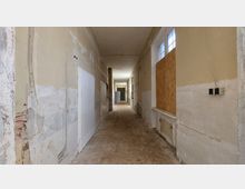 Ein leerer, schmaler Flur mit abgenutzten Wänden und Betonboden, an dem weiße Türen und zugemauerte oder mit Holzplatten abgedeckte Fenster zu sehen sind. Die Decke ist schlicht, und die Beleuchtung erfolgt durch natürliches Licht, das durch einige Fenster einfällt.