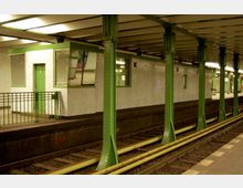 Ein U-Bahnhof mit grün gestrichenen Metallstützen, weiß gekachelten Wänden und gelben Schienenlinien. Im Vordergrund ist ein kleines Gebäude mit einer grünen Tür und großen Fenstern erkennbar.