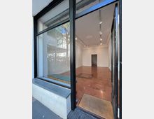 Ein leerer Verkaufsraum mit großen Fenstern und Glastür, der einen Blick nach innen ermöglicht. Der Raum hat einen hellbraunen Fliesenboden, weiße Wände, Stuckornamente an der Decke und eine schlichte Beleuchtung mit Schienensystem.