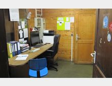 Ein kleiner Büroraum mit Holzwänden, einem Schreibtisch mit Computer, Monitor und Büromaterial sowie einem schwarzen Bürostuhl. An der Wand befinden sich ein grünes Pinnboard, ein Drucker und Regale mit Aktenordnern.