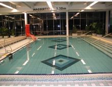 Hallenbad mit rechteckigem Becken, Fliesenboden in Blau- und Grüntönen mit diamantförmigen Mustern, 1,30 m Wassertiefe, breiter Einstieg mit Geländer und roter Rutsche am Beckenrand. Große Fensterfront mit Pflanzen im Hintergrund.