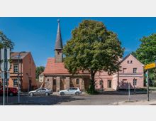 Historische Kirche aus Backstein mit spitzem, schindelgedecktem Turm, umgeben von Bäumen und angrenzenden hellrosa Gebäuden in einer städtischen Umgebung mit parkenden Autos und Straßenbeschilderung.