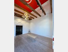 Heller, nahezu leerer Innenraum mit breiten Holzdielen, weiß verputzten Wänden und sichtbaren Holzbalken unter einer roten Deckenfläche. Ein farbiger Kronleuchter hängt von der Decke, dazu gibt es eine kleine Bank in der Mitte und einen Durchgang mit dekorierter Stuckumrahmung links.