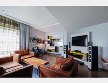 Wohnzimmer mit moderner Einrichtung, darunter braune Ledersofas, ein Couchtisch aus Holz, ein großer Flachbildfernseher an der Wand, Standlautsprecher und ein Bücherregal mit grünen und schwarzen Akzenten. Helle Vorhänge und eine Stehlampe sorgen für eine gemütliche Atmosphäre.