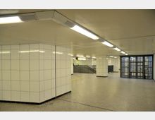 U-Bahn-Station mit einfacher Architektur, weißen gefliesten Wänden, grauem Boden und Deckenbeleuchtung. Sichtbare Treppen und Geländer führen in andere Bereiche der Station, im Hintergrund sind Glastüren und ein gelber Hinweis signalisierend.