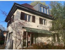 Zweistöckiges Wohnhaus mit weißer Fassade, braunen Fensterläden und einem Satteldach. Im Vordergrund sind ein kleiner Garten mit blühenden Sträuchern sowie eine überdachte Terrasse zu sehen.