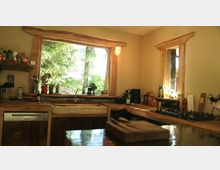 Rustikale Küche mit Arbeitsflächen und Fensterrahmen aus naturbelassenem Holz. Durch zwei große Fenster fällt Tageslicht und bietet einen Blick auf eine grüne, bewaldete Umgebung; auf den Arbeitsflächen stehen Küchenutensilien, Flaschen und Kochgeräte.