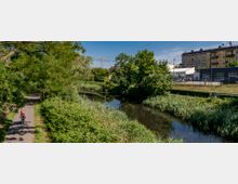 Ein schmaler Fluss mit dicht bewachsenen Ufern fließt durch eine grüne Parklandschaft. Rechts davon stehen moderne, flache Gebäude und ein gelbes Mehrfamilienhaus, daneben verläuft ein Radweg mit einem Radfahrer.