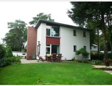 Zweistöckiges Einfamilienhaus mit weißer Putzfassade und roten Akzenten, umgeben von einem gepflegten Garten. Im Vordergrund befindet sich eine Terrasse mit Gartenmöbeln, während im Hintergrund Bäume und Grünflächen zu sehen sind.