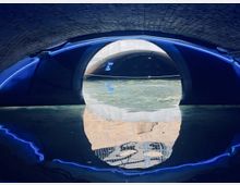 Unterirdischer Tunnel mit Wasser, beleuchtet durch blaue Neonlichter, die einen gekrümmten Bogen und die Struktur der gemauerten Decke betonen. Das Wasser reflektiert Teile der Tunnelbeleuchtung und der Umgebung.