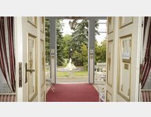 Blick durch eine geöffnete, verzierte Tür mit goldenen Details auf eine Terrasse mit rotbraunem Teppich und weißen Möbeln. Im Hintergrund eine Skulptur auf einem kleinen Platz, umgeben von Bäumen und Grünflächen.