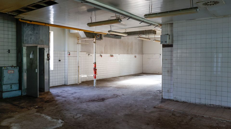 Leerer, stillgelegter Industrie- oder Küchenraum mit weiß gekachelten Wänden, Betonboden und einer abgehängten Decke mit Leuchtstoffröhren. Sichtbare Rohre, ein Metalltürbereich und wenige Fenster sorgen für spärliches Tageslicht.