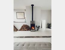 Ein minimalistischer Wohnraum mit einem Kaminofen in der Mitte, der ein Feuer brennt. Daneben ein Stapel Holzscheite, eine Topfpflanze und ein bodenlanger Spiegel; der Raum ist in neutralen Farben mit einfacher Dekoration gehalten.