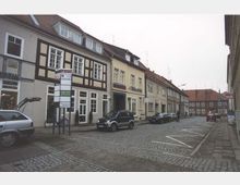 Eine Kopfsteinpflasterstraße in einer Altstadt mit zweigeschossigen Gebäuden im traditionellen Stil, darunter ein Fachwerkhaus. Am Straßenrand sind parkende Autos und ein Wegweiser mit Geschäftsinformationen sichtbar.