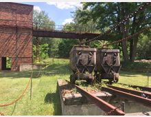 Backstein-Gebäude eines historischen Industriekomplexes mit angrenzenden Schienen und zwei verrosteten Förderwagen. Im Hintergrund ist eine grüne Waldlandschaft, sowie eine rostige Kettenkonstruktion erkennbar.