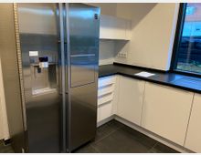 Eine moderne Küche mit weißen Schränken, einer schwarzen Arbeitsplatte und einem großen, silbernen Side-by-Side-Kühlschrank mit integriertem Wasser- und Eisspender. Rechts befindet sich ein großes Fenster, durch das Tageslicht einfällt.