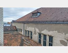 Blick auf die Dachlandschaft mehrerer Gebäude mit roten Ziegeln, darunter ein großes Gebäude mit sichtbaren Putzschäden an der Fassade und mehreren großen Fenstern. Im Hintergrund sind weitere Gebäude und ein bewölkter Himmel zu sehen.
