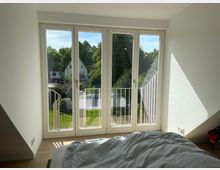 Helles, schlichtes Schlafzimmer mit großen bodentiefen Fenstertüren und kleinem Balkon mit weißem Geländer. Durch die Fenster blickt man auf einen grünen Garten mit Bäumen und benachbarte Häuser.