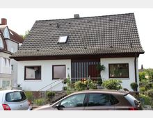 Ein Einfamilienhaus mit Satteldach, weißer Fassade und braunen Fensterrahmen. Der Eingangsbereich ist durch eine kleine Treppe mit Geländer erreichbar und von Pflanzen und Blumen umgeben; im Vordergrund parken mehrere Autos.