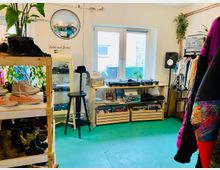 Ein kleiner Secondhand-Laden mit türkisfarbenem Boden, Regalen voller Schuhe, Kleidungsständern und einem Wandregal mit einem Plattenspieler. Im Hintergrund befindet sich ein Fenster, und Pflanzen sowie Dekorationsgegenstände sind im Raum verteilt.