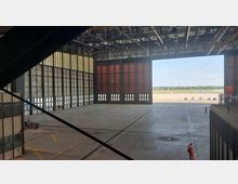 Großer Flugzeughangar mit hohen, teilweise offenen Toren, die den Blick auf das Außengelände eines Flughafens mit Rollfeld und einigen geparkten Fahrzeugen freigeben. Die Konstruktion besteht aus Stahlträgern, und das Innere zeigt eine leere, weitläufige Betonfläche.
