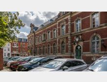 Straßenszene mit einem historischen Backsteingebäude mit verzierten Fenstern und grüner Eingangstür im Vordergrund; mehrere parkende Autos auf einem Kopfsteinpflasterplatz sowie weitere Gebäude im Hintergrund.