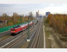 Zugstrecke mit einem roten Regionalzug der Deutschen Bahn, umgeben von herbstlicher Vegetation und einem Mix aus städtischen und industriellen Gebäuden im Hintergrund. Mehrere Gleise verlaufen parallel, gesäumt von Oberleitungen und Signalanlagen.