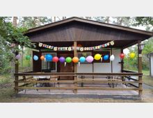 Eine kleine Holzhütte mit überdachter Terrasse, dekoriert mit bunten Luftballons und einer Girlande mit der Aufschrift „Alles Gute zum Schulanfang“. Im Hintergrund ist ein bewaldeter Außenbereich erkennbar.