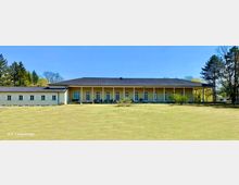 Ein einstöckiges, langes Gebäude mit heller Fassade und dunklem Satteldach, umgeben von einer offenen Wiese und Bäumen. Die Vorderseite des Gebäudes verfügt über große Fenster und Türen, die symmetrisch angeordnet sind.