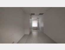 Leerer Raum mit weißen Fliesenboden und weißen Wänden, teilweise mit Fliesenspiegel. Ein Fenster mit vertikaler Jalousie und ein Heizkörper befinden sich am Ende des Raumes.