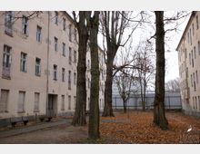 Ein Innenhof mit kahlen Bäumen inmitten mehrstöckiger, verputzter Wohngebäude mit verblasster Fassade und teils beschädigten Fenstern. Der Boden ist mit Laub bedeckt, und im Hintergrund ist ein Metallzaun sichtbar.