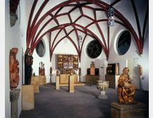 Innenraum eines Museums mit gotischem Gewölbe aus roten Backsteinbögen, weißen Wänden und runden Fenstern. Ausgestellt sind mittelalterliche Holzskulpturen und Altäre, dazu ein verzierter Taufstein in der Mitte.