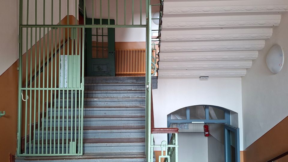 Treppenhaus in einem älteren Mehrfamilienhaus mit grauen Stufen, grünem Metallgeländer und einem Gittertor am Treppenaufgang. Sichtbar sind weiße Deckenbalken, ein bogenförmiger Durchgang mit blauem Türrahmen und zweifarbige Wände in Weiß und Beige.