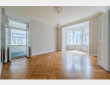 Heller, leerer Altbau-Wohnraum mit Fischgrätparkett, hohen Decken mit Stuck und weißer Pendelleuchte. Große Erkerfenster mit Heizkörpern darunter sowie Flügeltüren führen in einen angrenzenden Raum mit Einbauschränken.