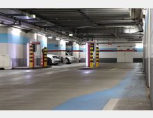 Unterirdischer Parkplatz mit beleuchteten Fahrspuren, Schranken und gelb-schwarz gestreiften Sicherheitspfosten. Im Hintergrund sind parkende Autos, blau-weiß gestrichene Wände und Lüftungsrohre zu sehen.