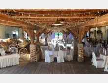 Ein rustikaler Veranstaltungsraum in einer Scheune mit Holzbalkendecke und freiliegenden Ziegelpfeilern, dekoriert mit Lichterketten. Der Raum ist mit runden Tischen, weißen Stuhlhussen und Kerzen ausgestattet; im Hintergrund steht ein mit Stroh gefüllter Karren.