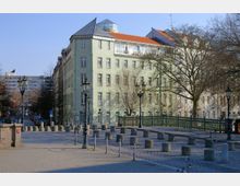 Mehrstöckiges, hellgrünes Wohn- oder Bürogebäude mit Spitzdach und Dachfenster, umgeben von kahlen Bäumen. Im Vordergrund eine gepflasterte Straße mit Pollern, dekorativen Straßenlaternen und einer grünen Metallbrücke.