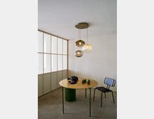 Minimalistisch eingerichteter Raum mit einem runden Tisch aus Holz mit grünem Sockel, darauf liegen Artischocken und Kopfhörer. Im Hintergrund eine halbhohe Milchglas-Wand, daneben eine schwarze, schlichte Metallstuhllehne, und von der Decke hängt eine mehrflammige Hängelampe mit Glaskugeln.