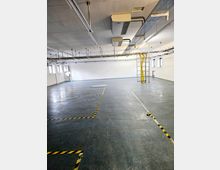 Großer, leerer Raum mit grauem Boden, weißen Wänden und einer vertäfelten Decke mit Lüftungssystemen und Kabeltrassen. Gelb-schwarze Markierungen am Boden zeigen abgegrenzte Bereiche, und eine Leiter steht an der Wand.