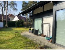 Einfamilienhaus mit moderner Fassade aus weißen Ziegeln und dunklem Holz, angrenzend an einen gestalteten Garten mit Rasenfläche und hohen Bäumen. Im Vordergrund eine gepflasterte Terrasse mit Gartenmöbeln und Blick auf Nachbarhäuser mit roten Ziegeldächern.