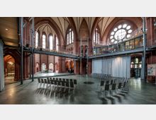 Innenraum einer historischen Backsteinkirche, umgebaut zu einem modernen Veranstaltungsraum mit offenen Emporen und Glaselementen. Der Raum hat hohe Spitzbogenfenster, eine Rosette, freistehende Stuhlreihen und Vorhänge, die den Bereich flexibel abtrennen.