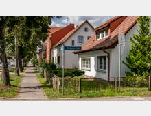 Reihenhäuser mit roten Ziegeldächern entlang einer baumgesäumten Straße, beschildert als „Wildparkstraße“, mit Gehweg und kleinen Vorgärten. Im Hintergrund sind parkende Autos und weitere Gebäude sichtbar.