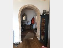 Innenansicht eines Wohnraums mit gemauertem Rundbogen, der in einen Nebenraum führt. Der Raum hat Holzboden, links steht ein gefülltes Hundenapfset, rechts ein blaues Regal mit Geschirr und Dekor. Im Hintergrund sind eine verzierte Holztruhe, ein Spiegel und Haken mit Jacken zu sehen.