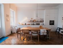 Bright open-plan kitchen and dining area with white built-in cabinets, a long countertop with sink and oven, and a large wooden table surrounded by mixed chairs. Tall windows on the left bring in natural light, and the room has a herringbone wood floor and a hanging pendant lamp over the table.