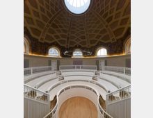 Ein runder Hörsaal mit weißen, halbkreisförmigen Sitzreihen und kunstvollen Geländern, darüber eine hohe Kuppeldecke mit geometrischem Muster und einem runden Oberlicht. Die Wände sind mit Fenstern und kunstvollen Wandmalereien verziert.