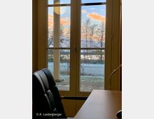 Blick durch eine verglaste Terrassentür auf eine schneebedeckte Terrasse, dahinter kahle Bäume und ein Zaun vor einem See mit Sonnenaufgang am Himmel. Im Vordergrund sieht man einen schwarzen Bürostuhl und einen Schreibtisch.