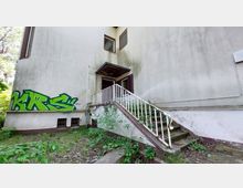 Außenansicht eines verwitterten Gebäudes mit weißer Fassade, grünen Graffiti-Buchstaben („KRS“) an der Wand und einer Metalltreppe, die zu einer Eingangstür mit Vordach führt. Vor dem Gebäude wachsen Pflanzen und Gras auf dem unbefestigten Boden.