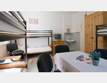 Ein einfacher Mehrbettzimmerraum mit Etagenbetten, einem Einzelbett, einem kleinen Holztisch und mehreren Stühlen. Im Hintergrund befinden sich ein kleiner Fernseher auf einer Kommode, ein Waschbecken an der Wand, ein Kleiderschrank und dekorative Details wie ein gerahmtes Bild und eine Pflanze auf dem Esstisch.