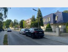 Eine ruhige Wohnstraße mit geparkten Autos entlang beider Seiten, rechts ein Wohnhaus mit blauem Dach und Vorgarten, links ein Gebäude mit Gerüst. Im Hintergrund sind Bäume und weitere Häuser zu sehen.